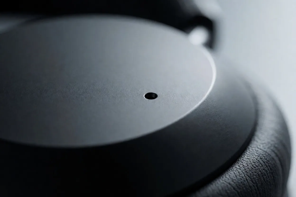 A close-up photograph of a black headphone earcup with a tiny pinhole microphone opening
