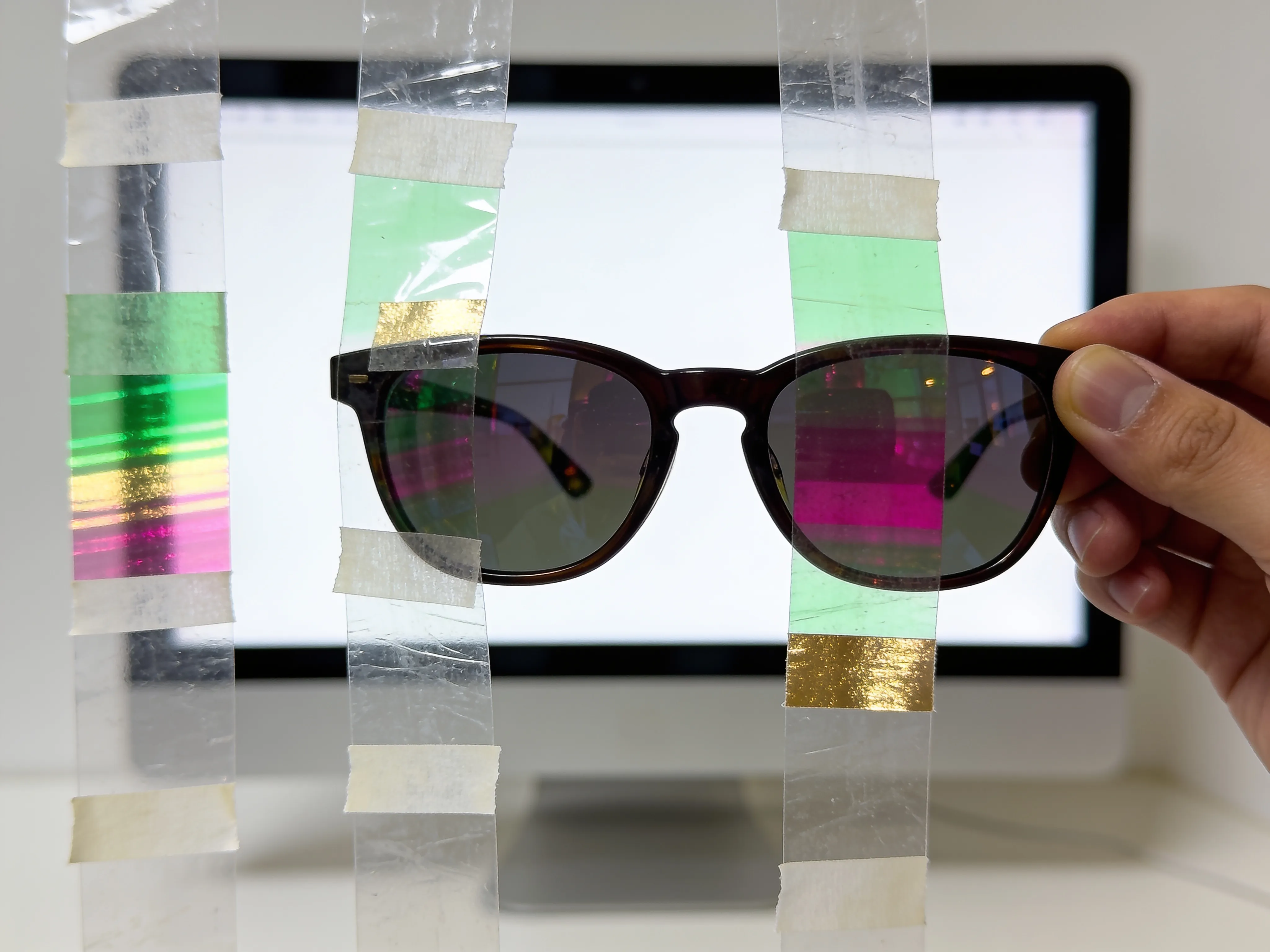 A photograph of clear tape strips on plastic, glowing green, magenta and gold, seen through sunglasses